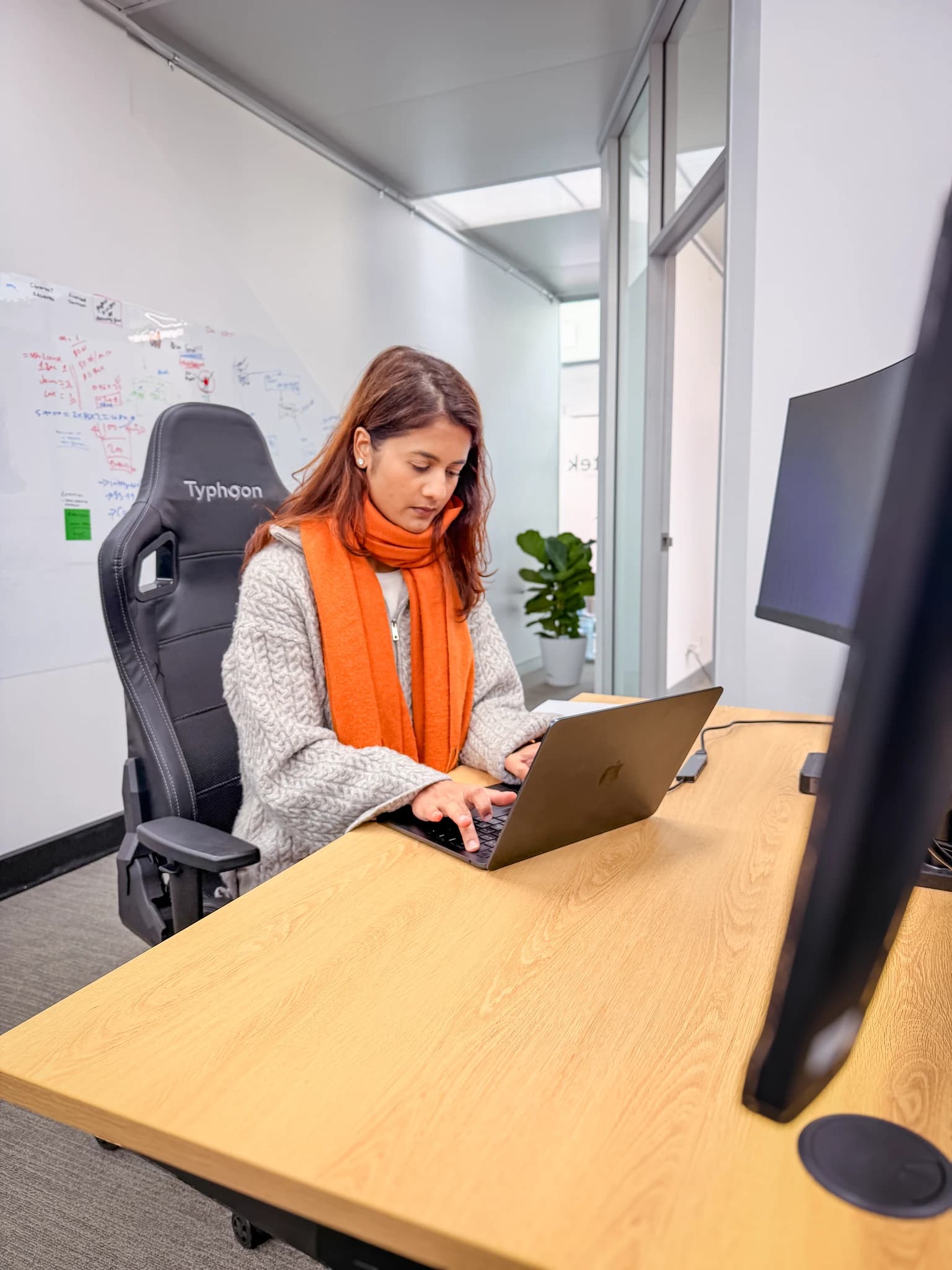 A woman wearing a grey sweater and a bright orange scarf sits in a black office chair, working on a dark laptop at a light wooden desk.